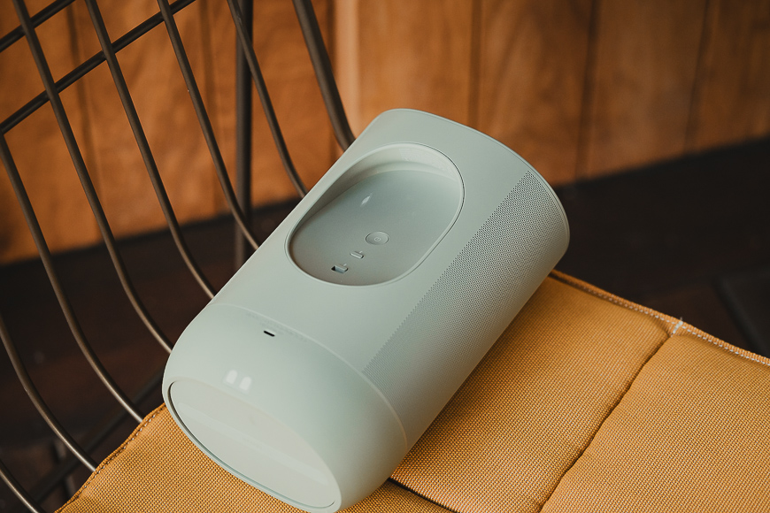 A cylindrical portable wireless speaker in light gray sits on a metal chair with a yellow cushion.
