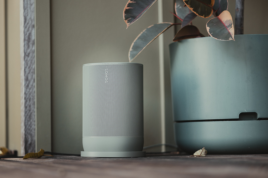 A light gray Sonos wireless speaker sits on a wooden surface next to a large green plant pot with a leafy plant.