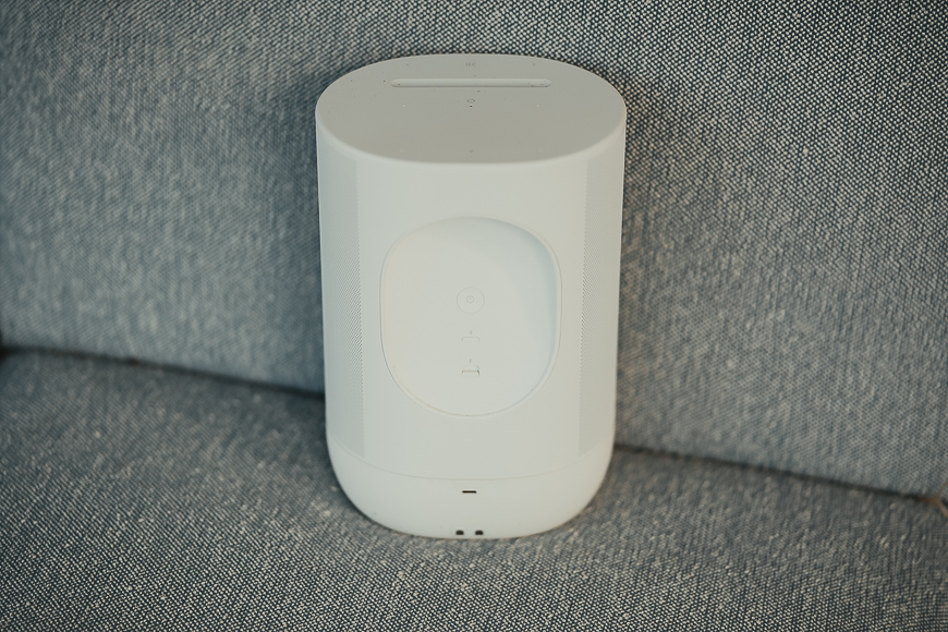 A white, cylindrical wireless speaker with buttons on the front sits on a gray fabric surface.