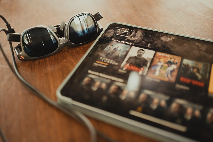 A pair of sunglasses and a tablet displaying a streaming service interface rest on a wooden surface.