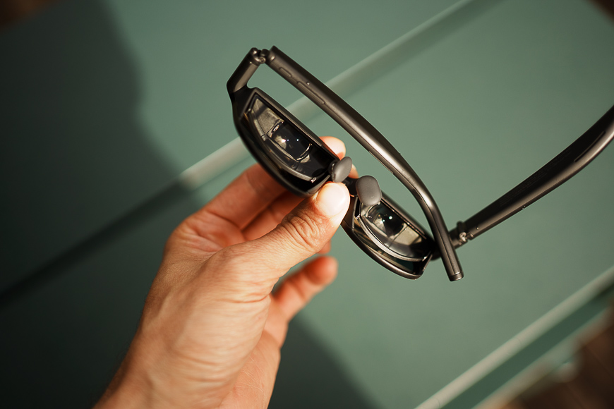 A hand holds a pair of black smart glasses with visible cameras and sensors on the inner side of the frames, above a green background.