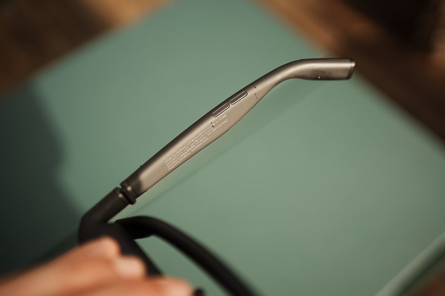Close-up of the arm of a pair of eyeglasses or sunglasses, showing two small buttons and engraved text on a matte metallic surface.