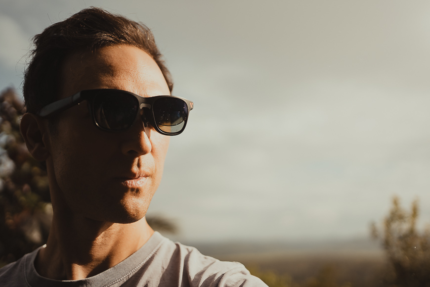 A man wearing dark sunglasses and a light shirt stands outdoors, looking to his left, with a blurred natural landscape in the background.