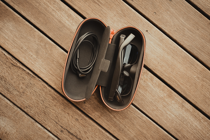 An orange sunglasses case lies open on a wooden surface, containing a pair of black sunglasses and a cable.