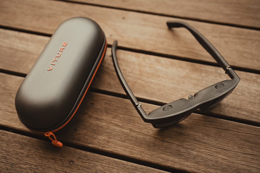 A pair of black smart glasses next to a closed, oblong Viture-branded carrying case on a wooden surface.