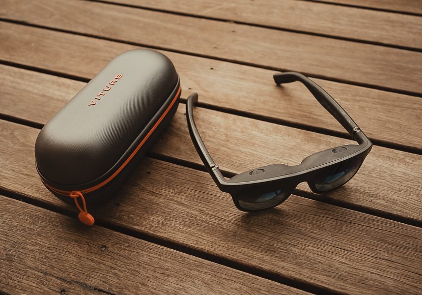 A pair of black smart glasses and a Viture-branded carrying case rest on a wooden surface. A pair of black smart glasses and a Viture-branded carrying case rest on a wooden surface.