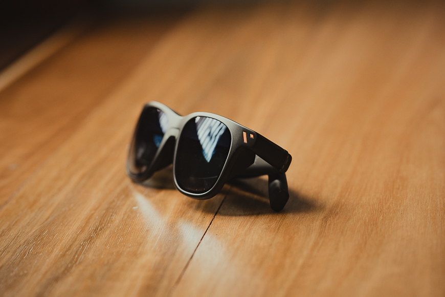 A pair of black sunglasses with thick frames rests on a wooden surface, reflecting light from a nearby window.