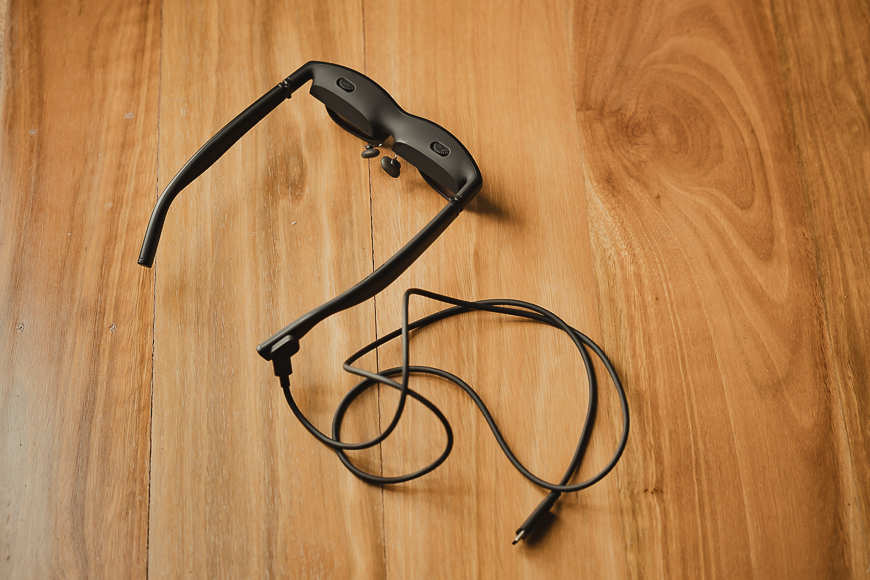 A pair of black smart glasses with a connected USB charging cable rests on a wooden surface.