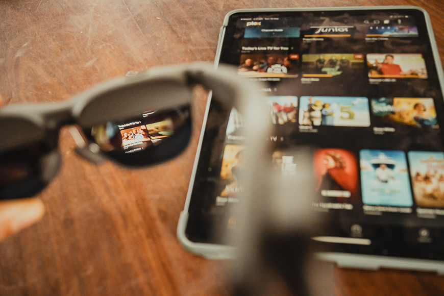 A person holds gray sunglasses in front of a tablet displaying a streaming service interface on a wooden surface.