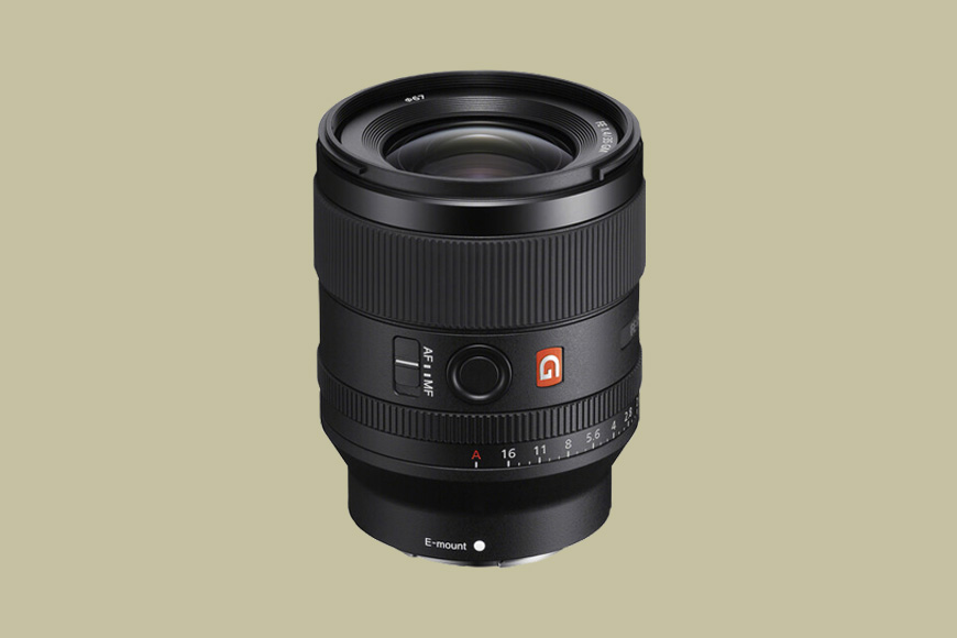 A black Sony G Master camera lens with manual focus and aperture control rings, displayed upright against a plain beige background.