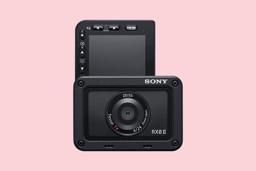 A Sony RX0 II camera with its LCD screen flipped up, set against a pink background. The lens has Zeiss branding and several control buttons are visible beside the LCD screen.
