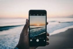 A person's hand holding an iphone capturing a photo of a seaside sunset.