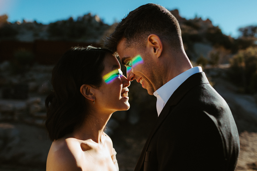 A couple standing close outdoors, facing each other and smiling. Sunlight creates rainbow reflections on their faces. The background is a rocky, sunlit landscape.