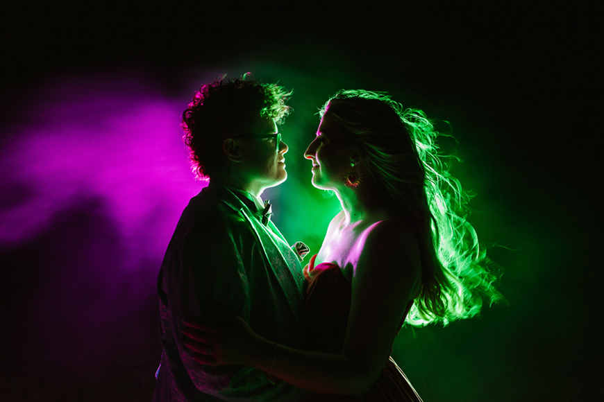 A couple stands close together in dim lighting, with vibrant pink and green backlighting creating a colorful aura around them.