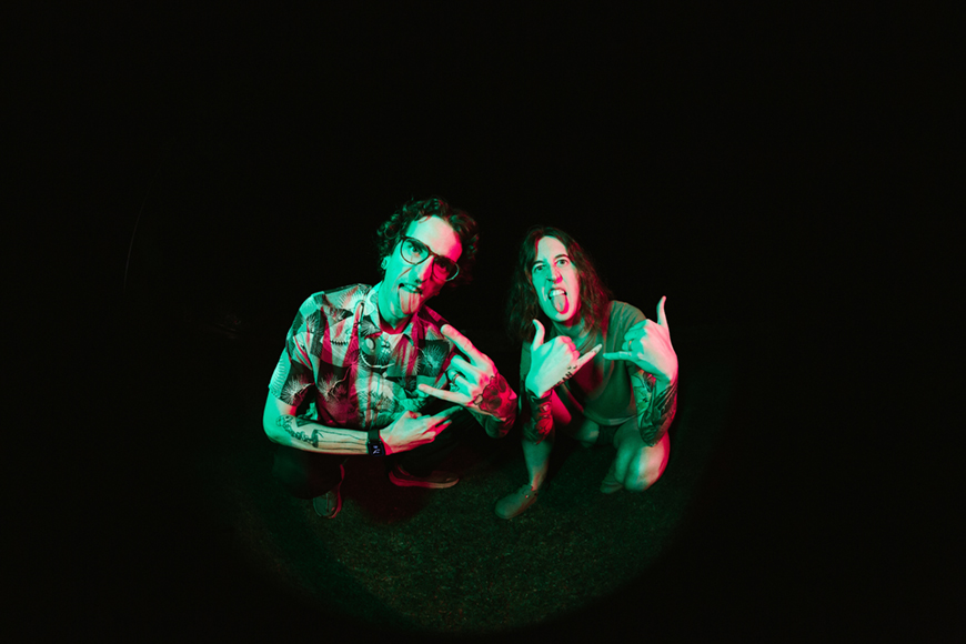 Two people posing with tongues out, displaying hand gestures under red and green lighting, against a dark background.