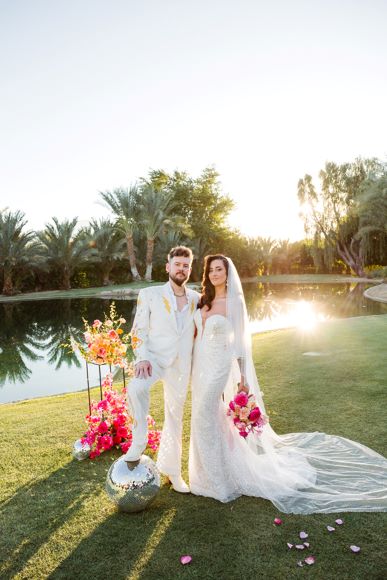 A couple in formal attire poses by a lake with a floral arrangement. The man is in a white suit, and the woman wears a white dress with a long veil. The sun is setting in the background.