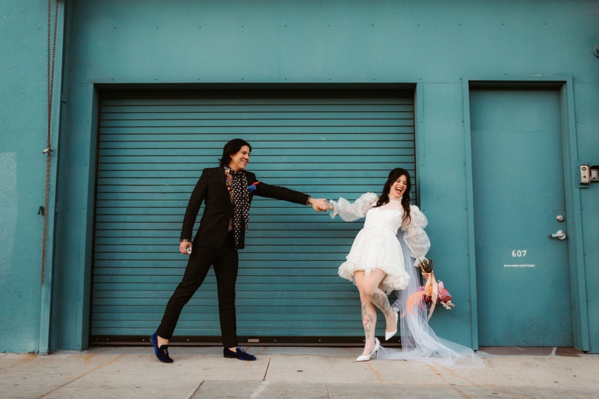 A couple stands in front of a blue roller door. The woman, in a white dress, holds flowers and is pulling the man, in a black suit, forward as they both smile.