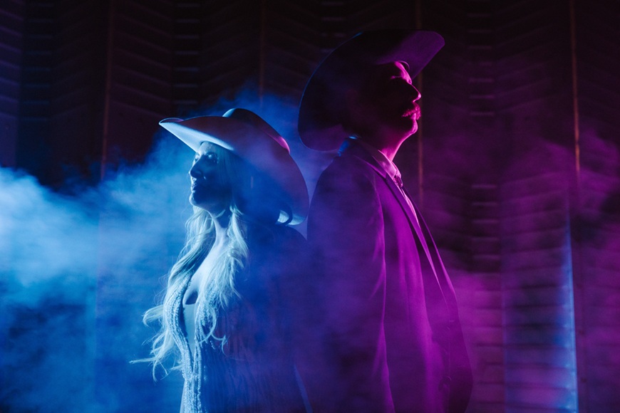Two individuals wearing cowboy hats stand back-to-back in smoke, with blue and pink lighting.