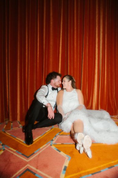 A man and woman in formal attire sit on a patterned carpet, leaning against an orange curtain, smiling at each other.