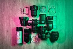 Photography equipment arranged on a wooden surface, including cameras, lenses, memory cards, camera accessories, and lighting gear, illuminated by green and pink lighting.