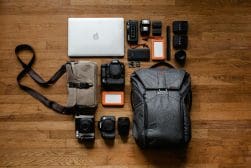 Photography equipment laid out on a wooden floor, including a laptop, cameras, lenses, memory cards, hard drives, camera straps, and a grey backpack.
