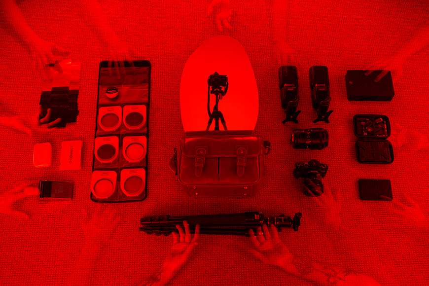Flat lay of camera gear including a camera, lenses, filters, a tripod, a camera bag, and accessories, all arranged neatly on a textured red background. Multiple ghosted hands are visible.