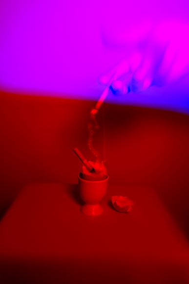 A hand holds a smoking incense stick over a small cup on a surface, illuminated by red and purple lighting.
