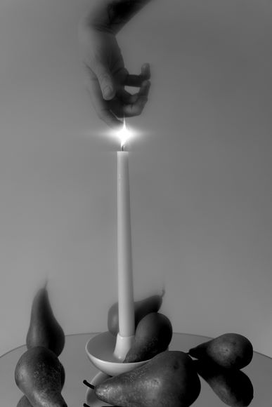 A hand hovers above a lit candle on a plate surrounded by pears, against a plain background.
